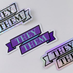 Pode incluir: Três adesivos com as palavras "THEY THEM" em um design de banner. Os adesivos têm um esquema de cores holográfico, roxo e azul. Os adesivos estão em um fundo branco.