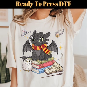 May include: Cream-colored t-shirt featuring a cartoon dragon wearing a scarf, glasses, and sitting on books. The design includes an owl, a golden snitch, and the text "Ready To Press DTF" at the top. The dragon is black with yellow and red accents.