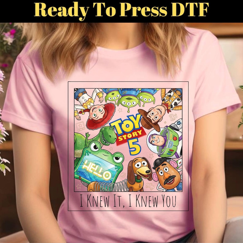 May include: A light pink t-shirt featuring a square graphic print of Toy Story characters including Woody, Buzz Lightyear, Jessie, Rex, Slinky Dog, Mr. Potato Head, and aliens. The text reads Toy Story 5 and I KNEW IT, I KNEW YOU.