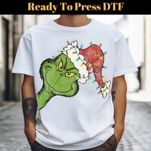 Op de afbeelding: Wit T-shirt met een groene Grinch-personage met een rode kerstmuts versierd met kleurrijke kerstverlichting. De tekst "Ready To Press DTF" staat bovenaan. De Grinch heeft een ondeugende uitdrukking.