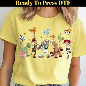 Puede incluir: Camiseta amarilla clara con un dise&ntilde;o de dibujos animados colorido de personajes animados, incluyendo un dinosaurio, un cerdo y un vaquero. El dise&ntilde;o incluye globos y estrellas. El texto "Ready To Press DTF" est&aacute; en la parte superior.