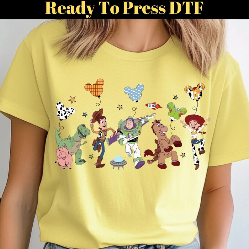 Puede incluir: Camiseta amarilla clara con un dise&ntilde;o de dibujos animados colorido de personajes animados, incluyendo un dinosaurio, un cerdo y un vaquero. El dise&ntilde;o incluye globos y estrellas. El texto "Ready To Press DTF" est&aacute; en la parte superior.