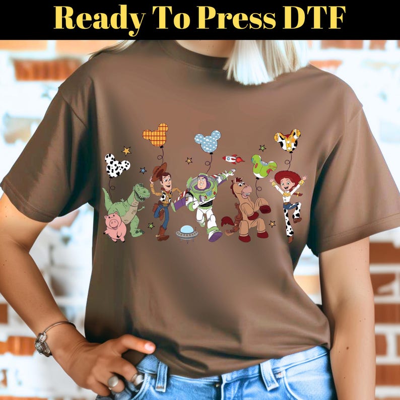Puede incluir: Camiseta marr&oacute;n con un gr&aacute;fico colorido de personajes animados con globos. El texto "Ready To Press DTF" est&aacute; en la parte superior. El dise&ntilde;o incluye un dinosaurio, un cerdo, un vaquero, un astronauta, un caballo y una vaquera.