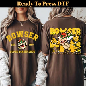 May include: Brown t-shirts featuring a Bowser graphic from Super Mario Bros. The front of the shirt displays "Bowser" in yellow lettering, with the character's image and "Est. 1985, Super Mario Bros." The back has a larger Bowser graphic.