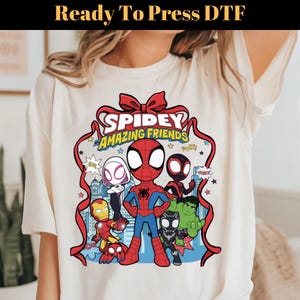 May include: Cream-colored t-shirt featuring a colorful graphic of Spidey and his Amazing Friends. The design includes Spider-Man, Iron Man, Hulk, and other characters. Text at the top reads "Ready To Press DTF".