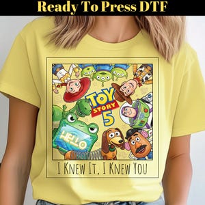 May include: A light yellow t-shirt featuring a square graphic print of Toy Story characters including Woody, Buzz Lightyear, Jessie, Rex, Slinky Dog, Mr. Potato Head, and Aliens. The text reads "TOY STORY 5" and "I KNEW IT, I KNEW YOU" with a frog holding a tablet.