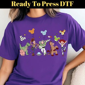 Puede incluir: Una camiseta morada con un gr&aacute;fico colorido de personajes animados, incluyendo un dinosaurio, un cerdo, un vaquero, un guardabosques espacial, un caballo y una vaquera. Los personajes sostienen globos. El texto "Ready To Press DTF" est&aacute; en la parte superior.