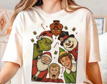 May include: A cream-colored t-shirt featuring a graphic print of various holiday characters including a green creature, Santa Claus, a snowman, an elf, a reindeer, and a young boy with hands on his face, surrounded by small gift boxes and candy canes.