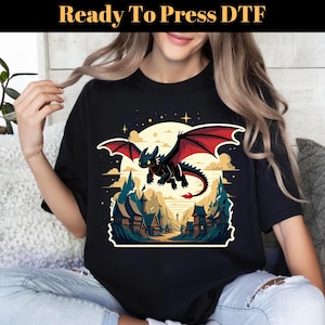 May include: Black t-shirt featuring a cartoon dragon with red wings, flying over a village scene. The design includes a full moon, stars, and the text "Ready To Press DTF" at the top. The dragon is black with red wings.