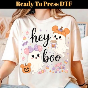 May include: A cream-colored t-shirt featuring a graphic print with the text "hey boo". The design includes two white ghosts, one wearing a purple bow and the other a pumpkin hat, surrounded by colorful candy corn, wrapped candies, and stars.