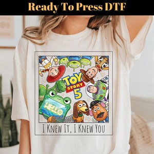 May include: A cream-colored t-shirt featuring a square graphic with Toy Story characters including Woody, Buzz Lightyear, Jessie, Rex, Slinky Dog, Mr. Potato Head, and Aliens. The text reads Toy Story 5 and I KNEW IT, I KNEW YOU.