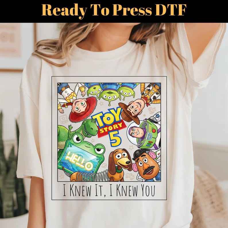 May include: A cream-colored t-shirt featuring a square graphic with Toy Story characters including Woody, Buzz Lightyear, Jessie, Rex, Slinky Dog, Mr. Potato Head, and Aliens. The text reads Toy Story 5 and I KNEW IT, I KNEW YOU.