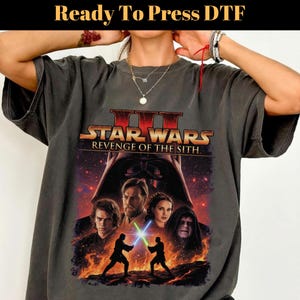Puede incluir: Una camiseta gris carbón con un estampado gráfico de Star Wars Revenge of the Sith. El diseño incluye el título de la película, el casco de Darth Vader y una escena central de dos figuras en duelo con sables de luz azul y verde.