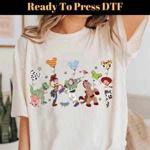 Puede incluir: Camiseta color crema con un gr&aacute;fico colorido de personajes animados, incluyendo un dinosaurio, un cerdo, un vaquero, un guardabosques espacial, un caballo y una vaquera. Los personajes sostienen globos. El texto "Ready To Press DTF" est&aacute; en la parte superior.