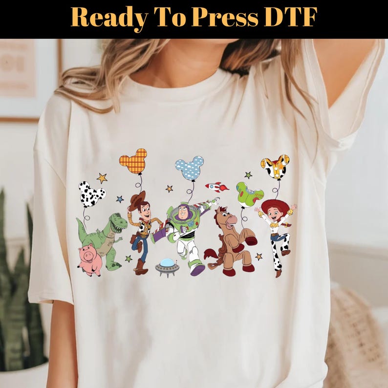 Puede incluir: Camiseta color crema con un gr&aacute;fico colorido de personajes animados, incluyendo un dinosaurio, un cerdo, un vaquero, un guardabosques espacial, un caballo y una vaquera. Los personajes sostienen globos. El texto "Ready To Press DTF" est&aacute; en la parte superior.