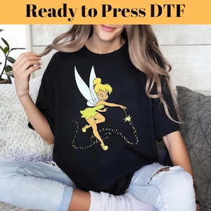 May include: A black t-shirt with a graphic of Tinkerbell, a fairy with yellow hair and a green dress, sprinkling fairy dust.