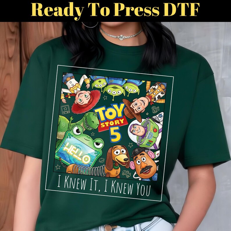 May include: A dark green t-shirt featuring a square graphic with Toy Story 5 characters including Woody, Buzz Lightyear, Jessie, Rex, Slinky Dog, Mr. Potato Head, and Aliens. The text reads Toy Story 5 and I KNEW IT, I KNEW YOU.