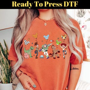Puede incluir: Camiseta naranja con estampado DTF (Direct to Film) de personajes de Toy Story con globos. El dise&ntilde;o incluye a Buzz Lightyear, Woody y Jessie. El texto "Ready To Press DTF" est&aacute; en la parte superior.