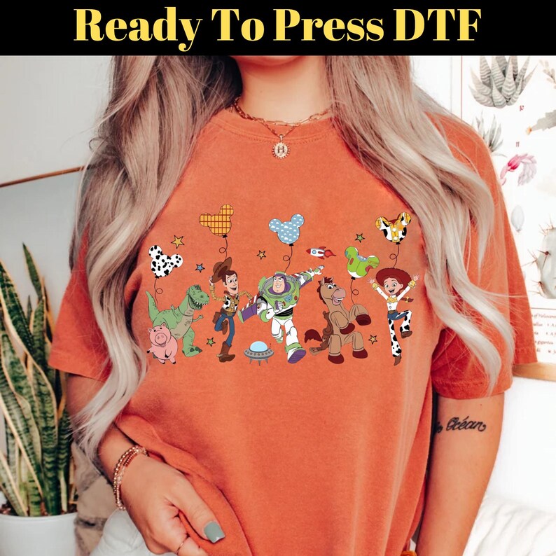 Puede incluir: Camiseta naranja con estampado DTF (Direct to Film) de personajes de Toy Story con globos. El dise&ntilde;o incluye a Buzz Lightyear, Woody y Jessie. El texto "Ready To Press DTF" est&aacute; en la parte superior.