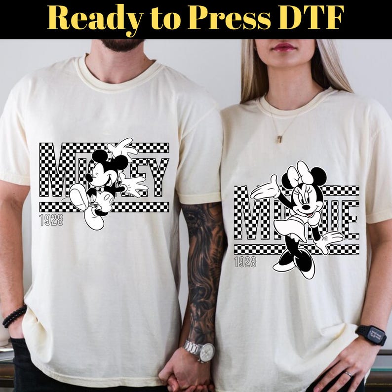 Op de afbeelding: Twee witte T-shirts met zwart-wit geruite ontwerpen met Mickey Mouse en Minnie Mouse. De shirts hebben de tekst "Mickey" en "Minnie" met het jaar "1928" eronder gedrukt.