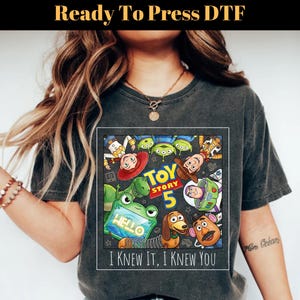 May include: A charcoal gray short-sleeve t-shirt featuring a square graphic print of Toy Story characters including Woody, Buzz Lightyear, Rex, and Slinky Dog. The text reads Toy Story 5 and I KNEW IT, I KNEW YOU. The design includes a green alien tablet.