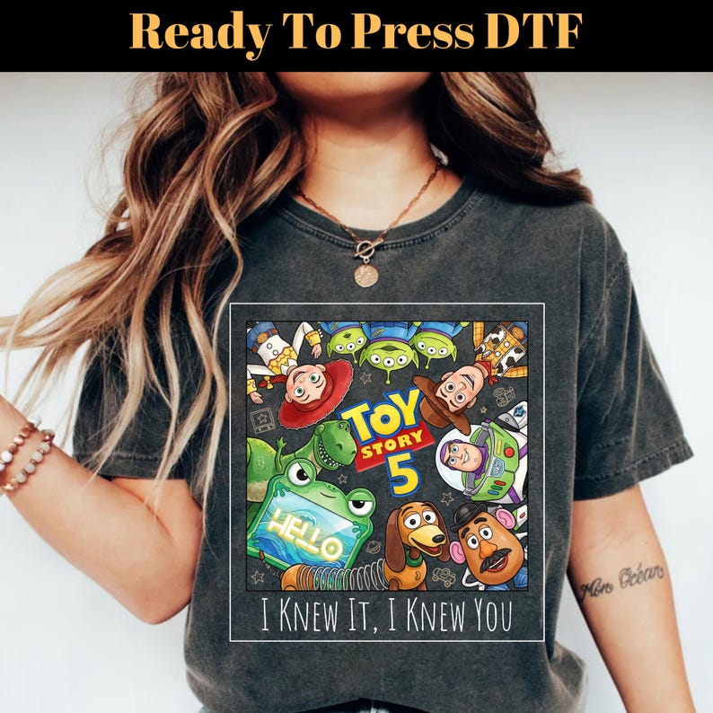 May include: A charcoal gray short-sleeve t-shirt featuring a square graphic print of Toy Story characters including Woody, Buzz Lightyear, Rex, and Slinky Dog. The text reads Toy Story 5 and I KNEW IT, I KNEW YOU. The design includes a green alien tablet.