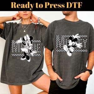Op de afbeelding: Twee grijze T-shirts met een zwart-wit geruit ontwerp met Mickey Mouse en Minnie Mouse. De shirts hebben de tekst "1928" erop gedrukt.