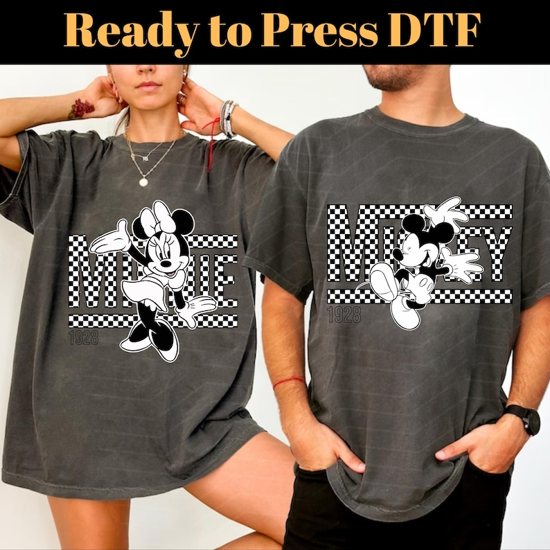 Op de afbeelding: Twee grijze T-shirts met een zwart-wit geruit ontwerp met Mickey Mouse en Minnie Mouse. De shirts hebben de tekst "1928" erop gedrukt.