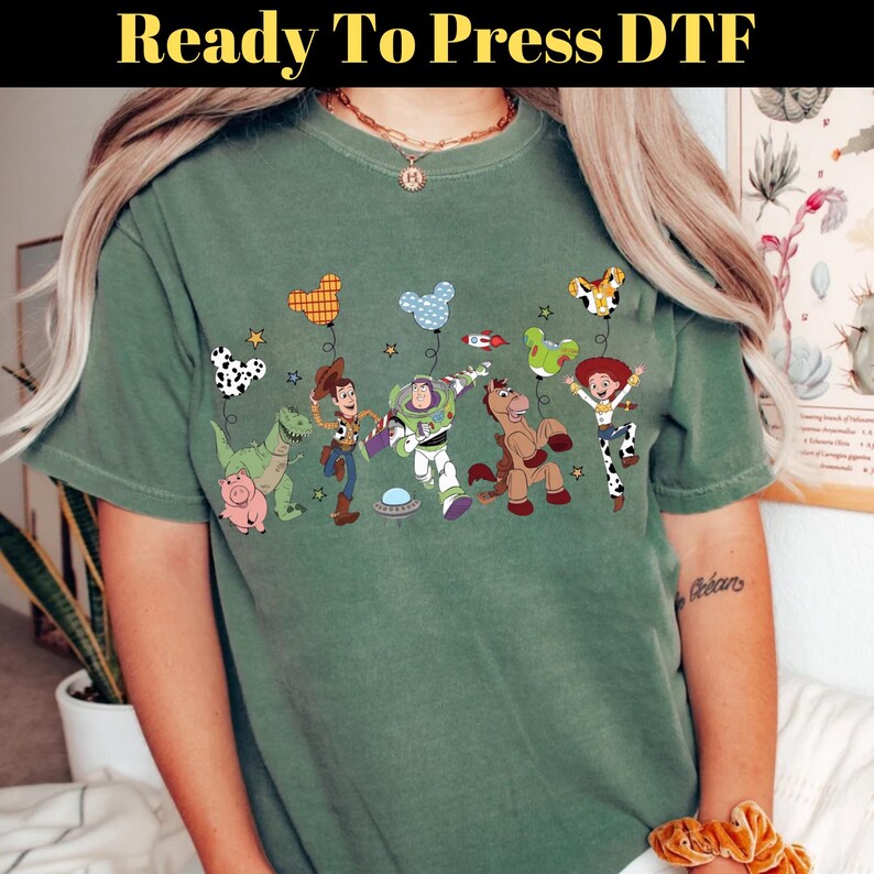 Puede incluir: Camiseta verde con un gr&aacute;fico colorido de personajes de dibujos animados, incluyendo un dinosaurio, un vaquero y un guardabosques espacial. El dise&ntilde;o incluye globos y estrellas. El texto "Ready To Press DTF" est&aacute; en la parte superior de la imagen.