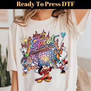 May include: Cream-colored t-shirt featuring a colorful graphic of Disney characters and the word "Epcot". The design includes Mickey Mouse, Minnie Mouse, Donald Duck, Goofy, and Pluto, with a futuristic cityscape in the background. The text "Ready To Press DTF" is at the top.
