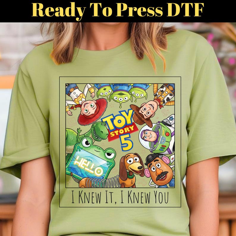 May include: A light olive green t-shirt featuring a square graphic print of Toy Story characters including Woody, Buzz Lightyear, Jessie, Rex, Slinky Dog, Mr. Potato Head, and aliens. The text reads Toy Story 5 and I KNEW IT, I KNEW YOU.
