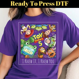 May include: A purple t-shirt featuring a graphic print of Toy Story characters including Woody, Buzz Lightyear, Jessie, Rex, Slinky Dog, Mr. Potato Head, and the Aliens. The text reads Toy Story 5 and I Knew It, I Knew You.
