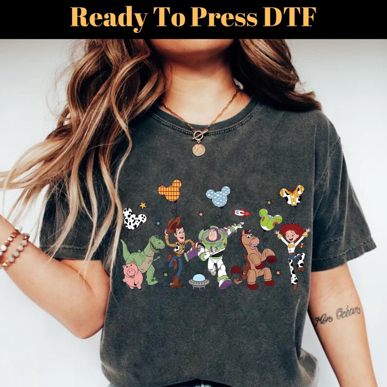 Puede incluir: Camiseta gris oscuro con un dise&ntilde;o colorido de personajes de Toy Story, incluyendo a Woody, Buzz Lightyear y Jessie. El dise&ntilde;o incluye globos y el texto "Ready To Press DTF". La camiseta es de estilo casual y c&oacute;modo.