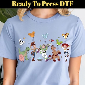 Puede incluir: Camiseta azul claro con un dise&ntilde;o colorido de personajes de dibujos animados y globos. El dise&ntilde;o incluye un dinosaurio, un cerdo, un vaquero, un astronauta, un caballo y una vaquera. El texto "Ready To Press DTF" est&aacute; en la parte superior.