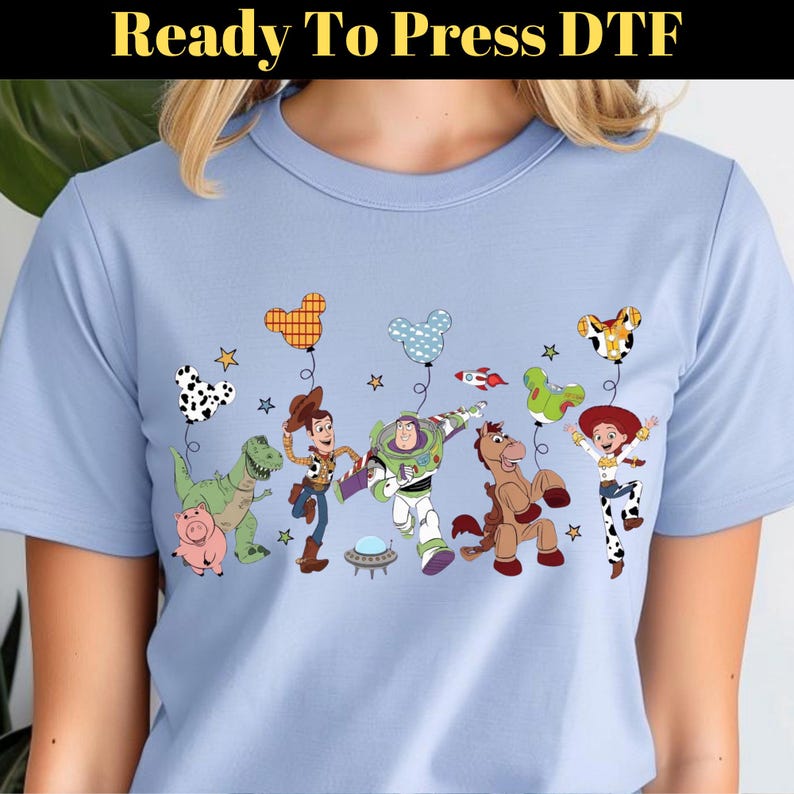Puede incluir: Camiseta azul claro con un dise&ntilde;o colorido de personajes de dibujos animados y globos. El dise&ntilde;o incluye un dinosaurio, un cerdo, un vaquero, un astronauta, un caballo y una vaquera. El texto "Ready To Press DTF" est&aacute; en la parte superior.
