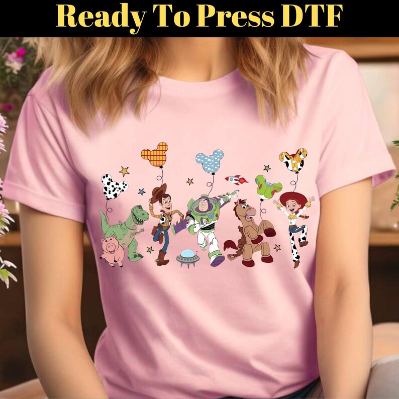 Puede incluir: Una camiseta rosa claro con un gr&aacute;fico colorido de personajes de dibujos animados. El dise&ntilde;o incluye globos, estrellas y el texto "Ready To Press DTF" en la parte superior.