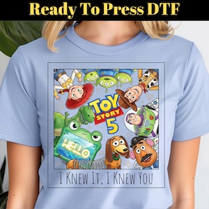 May include: A light blue t-shirt featuring a square graphic of Toy Story characters including Woody, Buzz Lightyear, Jessie, Rex, Slinky Dog, Mr. Potato Head, and Aliens. The text reads Toy Story 5 and I KNEW IT, I KNEW YOU.