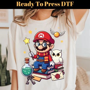 May include: A cream-colored t-shirt featuring a graphic of a cartoon character wearing a red hat with an M, glasses, and a striped scarf. The character holds a star wand while sitting on a stack of books next to an owl, a potion bottle, keys, and a letter.