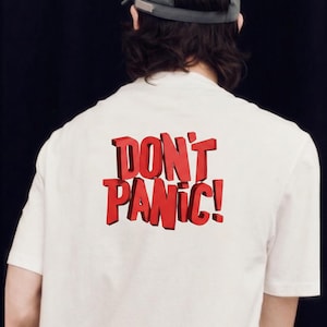 May include: A white short-sleeved t-shirt featuring the text DON'T PANIC! in bold, red, three-dimensional block lettering on the back. The shirt is worn with a grey baseball cap, viewed from the back against a plain black background.