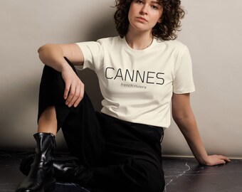 May include: A short-sleeved, off-white crew neck t-shirt featuring the black text CANNES in a bold, sans-serif font, with the words french riviera printed in a smaller, thinner font directly underneath the main text.