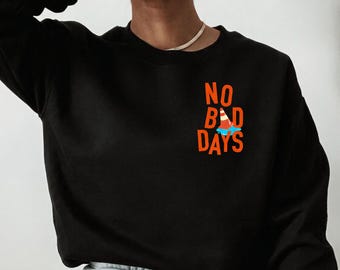 May include: A black long-sleeve sweatshirt featuring the orange text NO BAD DAYS printed on the left chest. The letter A in the word BAD is replaced by an orange and white traffic cone sitting in a small blue puddle of liquid.