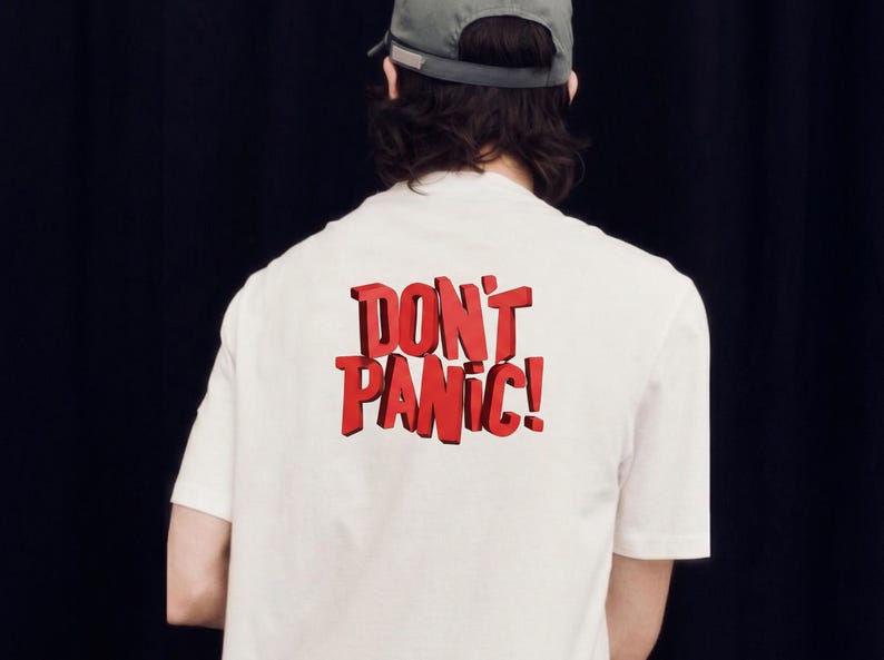 May include: A white short-sleeved t-shirt featuring the text DON'T PANIC! in bold, red, three-dimensional block lettering on the back. The shirt is worn with a grey baseball cap, viewed from the back against a plain black background.