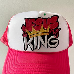 May include: A trucker hat with a white front panel and a pink brim and mesh back. The hat features the embroidered text "JESUS is my KING" in red and white, with a gold crown graphic.