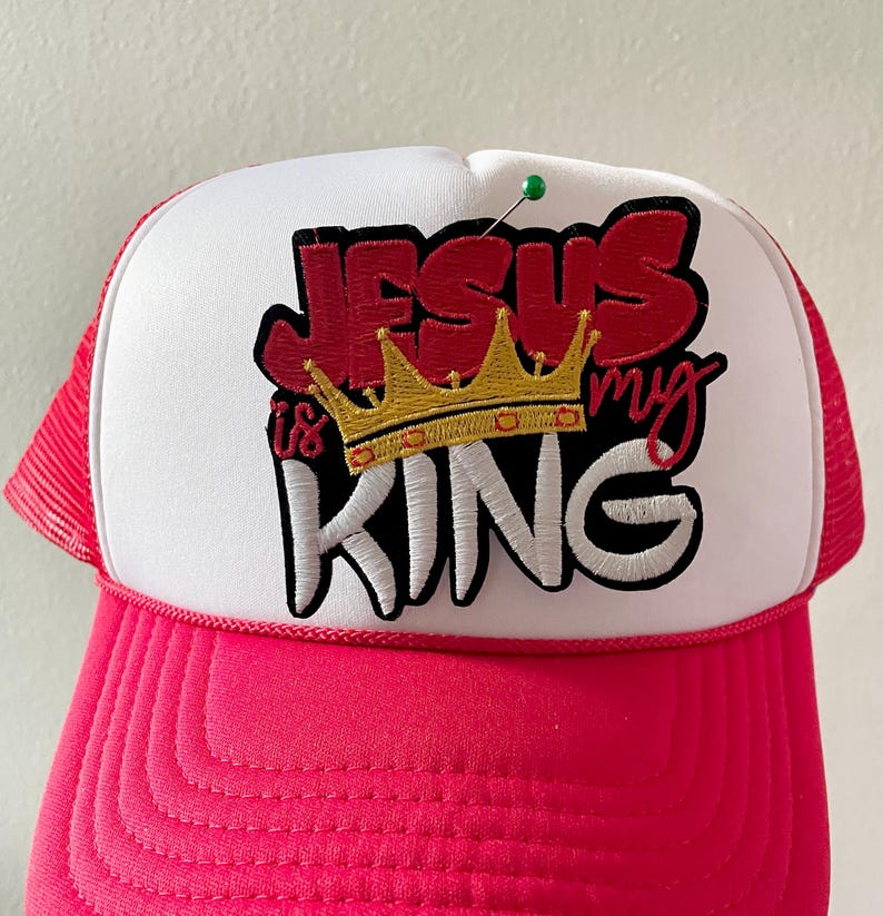May include: A trucker hat with a white front panel and a pink brim and mesh back. The hat features the embroidered text "JESUS is my KING" in red and white, with a gold crown graphic.
