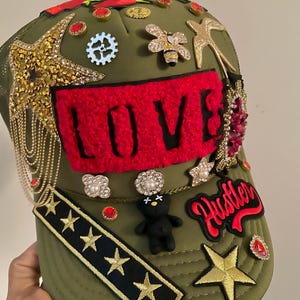 May include: A green trucker hat with a red "LOVE" patch, gold stars, a black teddy bear charm, and a red "Hustler" patch.