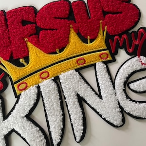 May include: Embroidered patch with the words "JESUS MY KING" in red and white, with a gold crown. The letters have a textured, fuzzy appearance, and the patch has a black border.
