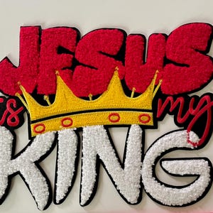 May include: Embroidered patch with the words "JESUS is my KING" in red, white, and black. A yellow and red crown is centered between "JESUS" and "KING". The patch has a textured, fuzzy appearance.