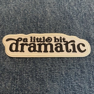 May include: A white embroidered patch with black lettering that says "a little bit dramatic".