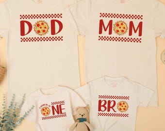 May include: Four white t-shirts and a onesie with pizza-themed designs. The shirts read "DAD", "MOM", and "BR", with a pizza graphic. The onesie says "CAMDEN IS ONE" with a pizza graphic. A small teddy bear is in the image.