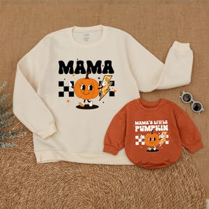 May include: A matching set of cream and orange sweatshirts with a cartoon pumpkin design. The adult sweatshirt says "Mama" and the child's sweatshirt says "Mama's Little Pumpkin".